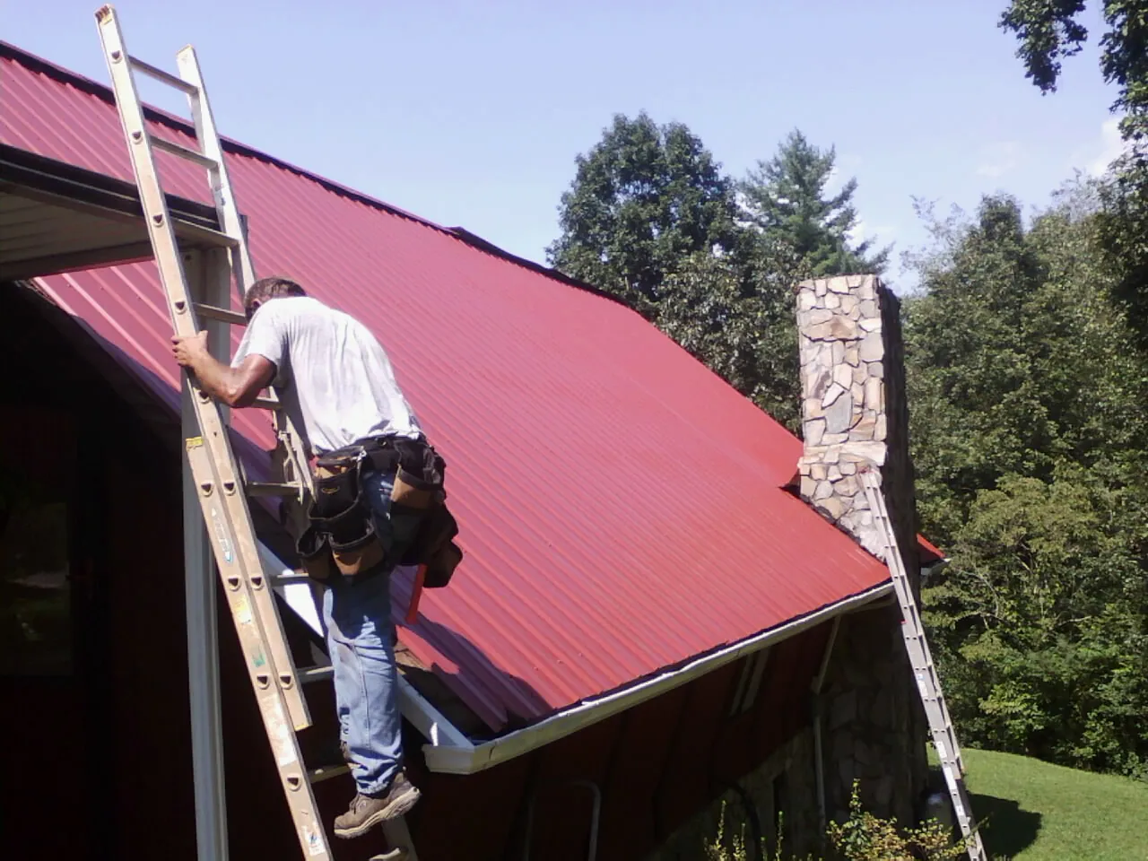 Roofer on a ladder beside a red metal roof and stone chimney — Soffit & Fascia Repair by Roofing Brothers in Saint Lucas
